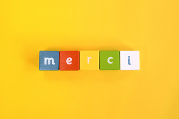 Merci - word made from colored wooden blocks. Painted cubes with letters on yellow background. Conceptual photo. Top view. Selective focus. Copy space.