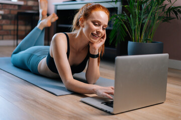 Sportive smiling redhead young woman lying on floor and using laptop computer at apartment. Sporty smiling female having online chatting via notebook while resting after sport training.