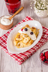 Rolled up thin pancakes stuffed with cottage cheese with berries and sour cream on a white wooden table.