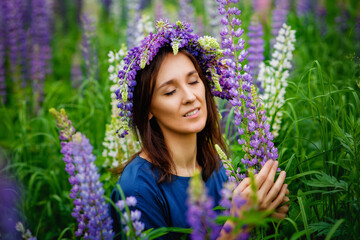A large portrait of a woman in a wreath on a lupine field with her eyes closed. A meadow of purple flowers in summer. A girl with long hair holds a bouquet of lupines against the background of nature.