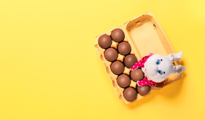 Package of ten chocolate eggs with fluffy white toy bunny on yellow background with copy space, empty text place. Banner for a religion holiday. Design mockup. Fun Easter hunt card. Handmade decor