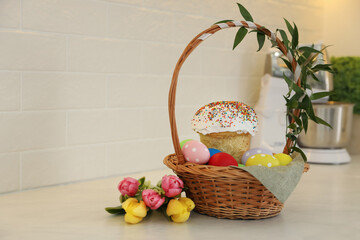 Fototapeta na wymiar Basket with traditional Easter cake, dyed eggs and flowers on table near white brick wall. Space for text