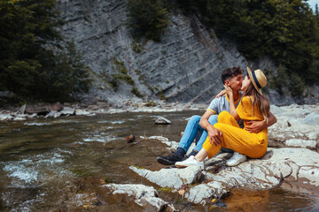 Couple of tourists kissing by mountain river enjoying landscape. Travelers sitting on rock. Summer vacation