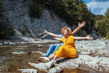 Couple of tourists relaxes by mountain river enjoying landscape. Travelers sit on rock with arms raised. Summer vacation