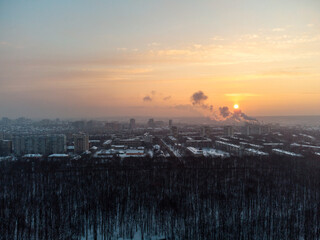 Aerial view Kharkiv city Pavlove Pole district. Smoke from pipes above winter dark forest, multistorey houses and snowy vibrant sunset sky