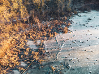 Aerial drone winter scenery on frozen wintery wild lake in forest with fallen trees in warm sunset light. Fly above natural park aerial view