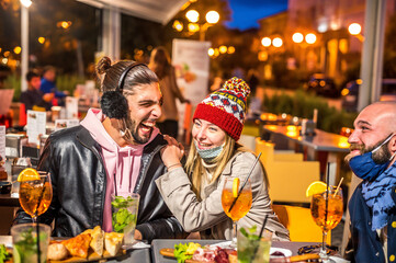 Couple drinking cocktail at bar in the night wearing face mask - New normal lifestyle concept with happy people friends having fun together toasting drinks at restaurant
