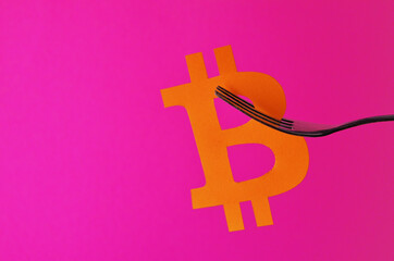 The orange bitcoin sign is hung on a fork on a pink background with a copy space. Conceptual collage about cryptocurrency in the style of minimalism. High quality photo