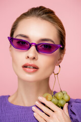 charming woman in purple eyeglasses, with fresh grapes earring isolated on pink