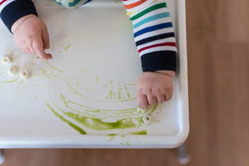 Young baby in rainbow stripe outfit spreading puff cereal and green puree on high chair tray; baby led weaning