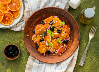 A healthy salad of sliced bloody oranges, purple onions, olives and pine nuts with olive oil dressing on a brown plate against an olive concrete background. Vegan recipes.