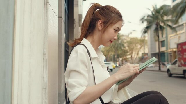 Successful young Asia businesswoman in fashion office clothes using digital tablet and typing text message while sitting alone outdoors in urban modern city in the morning. Business on the go concept.