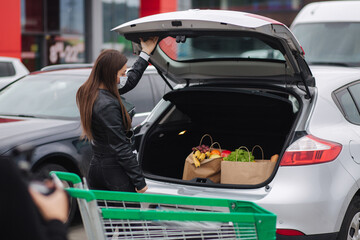 Young stylish woman in protective mask with shopping cart full of fresh food during quarantine, packing products into the car on the outdoor parking. Woman close car trunk on parking