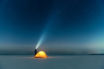 Tourist with flashlight near yellow tent lighted from the inside against the backdrop of incredible starry sky. Amazing night landscape. Tourists camp in snowy field. Travel concept - obrazy, fototapety, plakaty