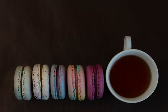 Small cakes makaroon with almonds and meringues of different colors lie near a cup with hot tea on a brown background