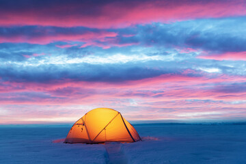Yellow tent lighted from the inside by a flashlight against the backdrop of glowind pink sky. Amazing snowy landscape. Tourists camp in winter field. Travel concept