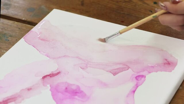 Close up of woman artist paints an abstract picture, she uses paintbrush to distribute water on canvas with pink paint