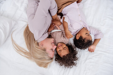 Multi ethnic happy family. Young blond Caucasian mother have fun with son and daughter African Americans on white bed. Woman hugs children kisses. Maternal care. Minimalist interior. Close up portrait