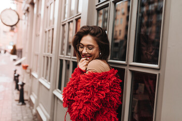 Coquettish woman in red knitted fluffy jacket smiles sweetly, shyly covering her mouth against...