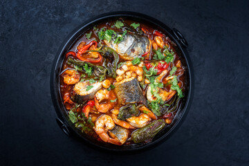 Modern style traditional Mexican seafood pozole soup with fish, king prawns and hominy in a clear sauce served as top view in design pot