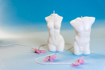 Isolated handmade soy wax candles on the blue background. Man and woman bodies made of a wax. Romantic shapes for candles. Close up on two aroma candles. Flowers and plants around the candles.