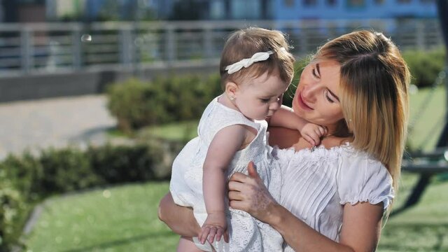 Cheerful blonde female holding adorable little daughter in her arms, lifting her up, kissing, hugging, gently throwing into air. Smiling loving mother enjoying outdoor leisure with cute toddler girl