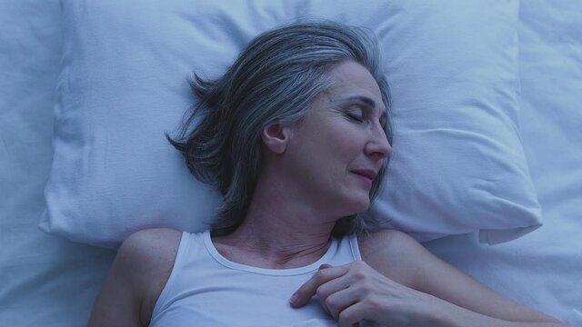 Mature lady sleeping in bed at white fresh linens, seeing sweet dreams, resting