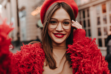 Young woman in bright red outfit and hat makes selfie. Lady in glasses with red lipstick posing against backdrop of beautiful building