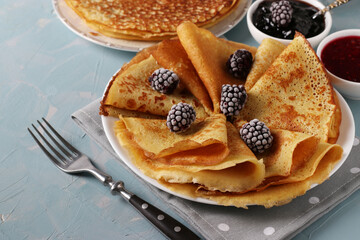 Homemade thin pancakes made from cornmeal, served with blackberries, jam and cup of milk on light blue background. Close-up. Horizontal format