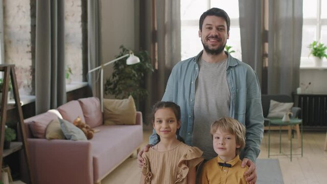 Slow-motion panning medium portrait of happy caucasian man and his two kids &ndash; son and daughter &ndash; standing in modern cozy living room smiling to camera