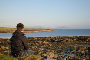 Fototapeta premium Young man wearing a black raincoat sat on the grass contemplating the coast with a low tide in the evening