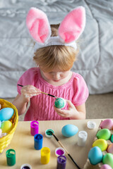 Easter holiday craft at home. Little girl in Easter bunny ears painting colored eggs.