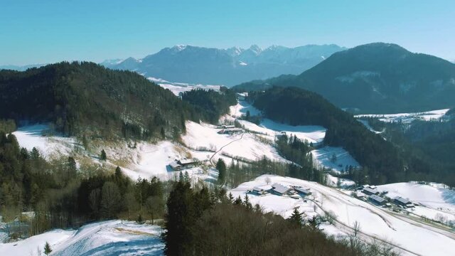 4K aerial drone flight moving above snowy hills and tree tops in the Bavarian Alps in winter in Germany, close to Austria. An alm chalet farm is seen on the way to the mountain peaks in the distance.