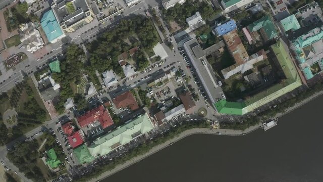 Aerial top view of a big city and embankment by the dark green river. Stock footage. Colorful roofs of houses and cars parked in the streets.