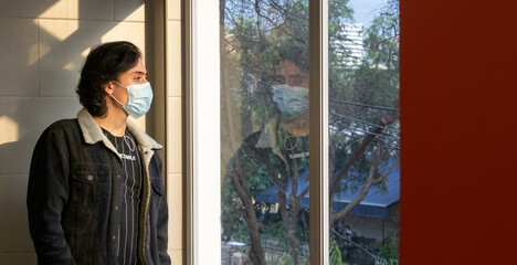 Young man looking out the window with mask due to quarantine caused by the covid 19 pandemic, concept of economic revival in the city, copy space