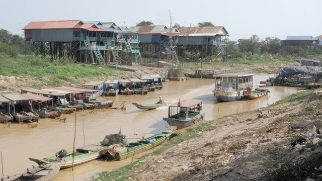 side view of a long tail fishing boat sailing through a floating village in the Tonle sap river during the dry season
