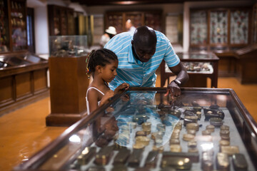 Afro male visitor and his little daughter visiting museum and looking at showcase with exhibits