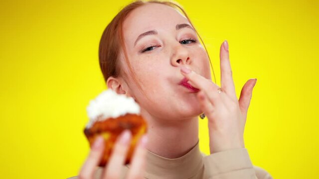 Close-up of funny green-eyed redhead Caucasian woman licking topping cream of fingers and smiling. Headshot portrait of joyful young slim lady enjoying delicious dessert at yellow background.