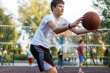 Cute young boy plays basketball on street playground in summer. Teenager in white t-shirt with orange basketball ball outside. Hobby, active lifestyle, sports activity for kids.