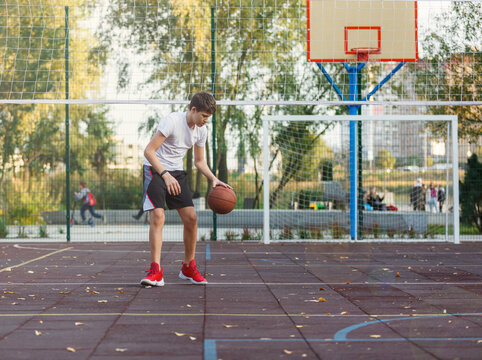 Cute Young Boy Plays Basketball On Street Playground In Summer. Teenager In White T-shirt With Orange Basketball Ball Outside. Hobby, Active Lifestyle, Sports Activity For Kids.