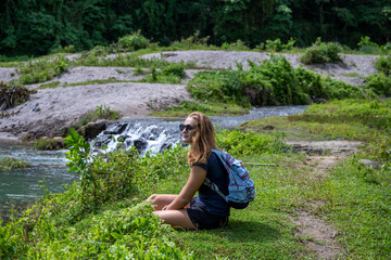 Relaxed female tourist sit on green grass near clean river. Green summer landscape. Candid woman portrait in natural environment. Hiking or trekking in forest. Tourism weekend activity