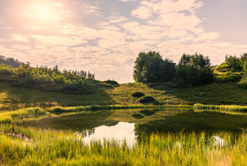 View of a mountain lake with smooth water and reflection, illuminated by the sun through the clouds.