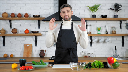 Smiling man chef in black apron looking camera tells teaches housewife records remote online video culinary webinar master class course in home kitchen, vegetables ingredients cooking salad on table