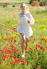 Naklejka na ściany i meble Cheery girl in white dress running in field with poppies plants