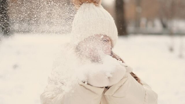 Winter holidays. Schoolgirl in the winter park. The kid in white jacket and hat blowing snow from hands.