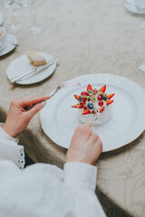 Photo of female hands cutting a fruit cake with strawberries, mint and black currants on a white plate on a table in a restaurant. dessert pavlova. table etiquette