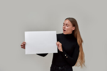 Naklejka premium Excited young caucasian woman with long hair dressed in black displaying and looking at white blank banner, standing isolated over gray background