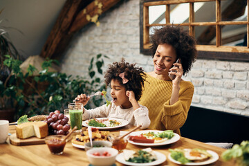 Happy black mother talking on the phone while having breakfast with her daughter in dining room.