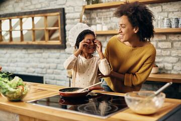 Happy African American mother and daughter having fun while cooking in the kitchen.
