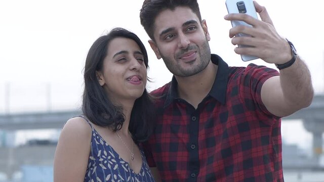 Beautiful young Indian couple taking selfies using smart phone while standing outdoors.Happy couple,friends making funny faces while taking selfies photos having fun together.